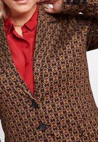 Mönstrad blazer i mörkbrun med ett geometriskt röd och gult mönster, med en spetsig krage och svarta knappar. Röd texturerad skjorta under.