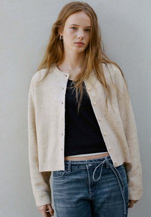 Junge Frau mit langen roten Haaren trägt einen beigefarbenen, geknöpften Cardigan über einem schwarzen Top und blauen Jeans, steht vor einem schlichten grauen Hintergrund.