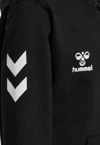 Sort stof med hvide dobbelte chevronstriber på ærmet og et bi-formet logo med teksten "hummel" på brystområdet.