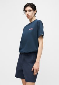 Νεαρός με κοντά σκουρόχρωμα μαλλιά που φοράει μπλε σκούρο t-shirt Ellesse και αντίστοιχα μπλε σκούρα σορτς, στέκεται μπροστά σε ουδέτερο φόντο.