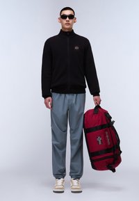 Felpa nera con zip, trousers grigi e sneakers beige. Tiene una borsa a tracolla rossa con una fascia nera e dettagli della bandiera norvegese.