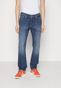 Denimjeans i mörkblått med rak passform, subtila blekningar och femficksdesign, bärs med orange sneakers med vita ränder.