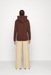 Person med flätat hår iförd en brun hoodie, beige vida byxor och vita sneakers, stående vänd mot en ljus enfärgad vägg.