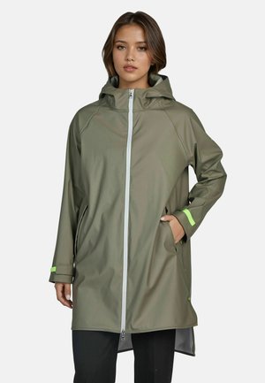 Junge Frau trägt eine olivgrüne Regenjacke mit Kapuze, vorn mit Reißverschluss und neongrünen Akzenten, steht mit einer Hand in der Tasche.