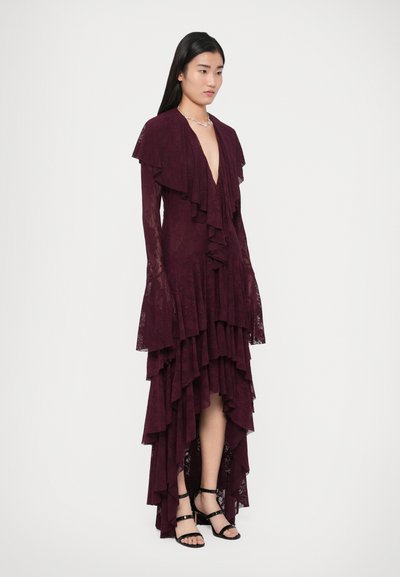 Abito di pizzo color mogano con dettagli arricciati, scollo a V profondo, maniche lunghe e orlo asimmetrico. Indossato con sandali neri con cinturini.