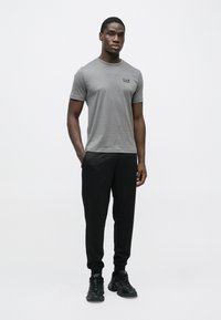 Pelēka īsroku T-krekls ar mazu melnu logotipu, kopā ar melnām šaurām biksēm un melniem sporta apaviem. Redzama gluda auduma tekstūra.