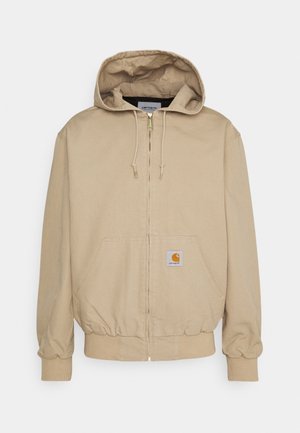Veste à capuche beige zippée avec poches avant, poignets et ourlet côtelés, et une petite écusson carré logo sur la poche avant.