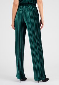 Pantaloni larghi in velluto verde con pieghe verticali, dotati di una vita elastica e una texture liscia. Scarpe col tacco nere visibili.