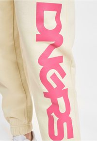 Beige Sweatpants mit großen, auffälligen pinken Buchstaben an der Seite. Elastische Bündchen an den Knöcheln, aus weichem, strukturiertem Stoff.