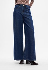 Jeans azul de perna larga feitos de denim, com uma cintura alta, fecho de botão e costura mínima. Usados com botas de salto pretas brilhantes.