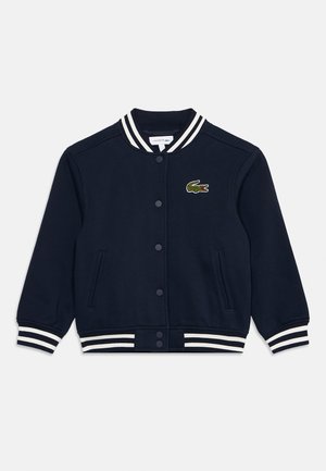 Marineblauwe varsityjas met knopen aan de voorkant, witte gestreepte manchetten en kraag, voorzakken en een klein groen krokodillenlogo op de borst.