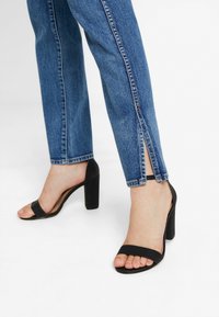 Sandales à talons noires avec une large bride sur le pied, design à bout ouvert et talon épais carré. Portées avec un jean bleu fendu sur les côtés.