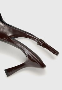 Ruskea nahkainen korkokenkä, jossa on kärjellinen muotoilu, pumps-design ja säädettävä hihna. Kiiltävä pinta ja minimalistinen siluetti.