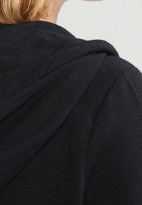 Blonde person wearing a black hooded sweatshirt, showing the back and side of the upper body and hood.