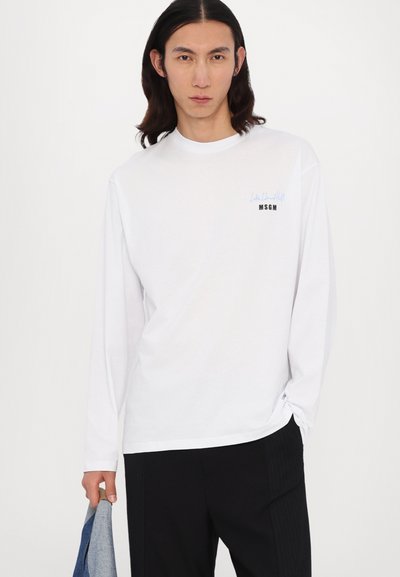 Tricou alb cu mânecă lungă și guler rotund. Prezintă text brodat în albastru și negru pe partea stângă a pieptului. Textura materialului este netedă.