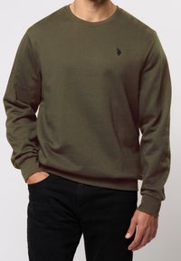 Oliivinvihreä pitkähihainen collegepaita, joka on valmistettu pehmeästä puuvillasta. Paidassa on pyöreä kaula-aukko ja huomaamaton musta logo yksityiskohtana rintamuksessa.