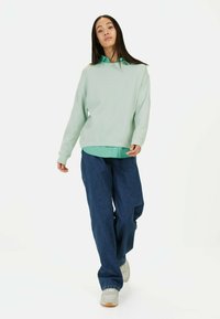 Lichtgroene ribbed sweatshirt over een blauwe shirt, gecombineerd met losse blauwe jeans en lichtgekleurde sneakers.