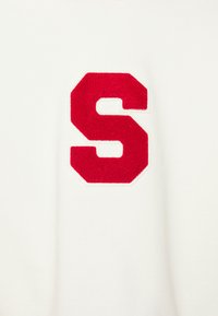 Ecusson texturé en forme de lettre "S" rouge, cousu sur un fond de tissu blanc uni.