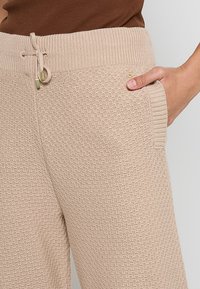 Beige knitted joggers with a textured pattern, elastic waistband, and a drawstring. Features a side pocket on the left.