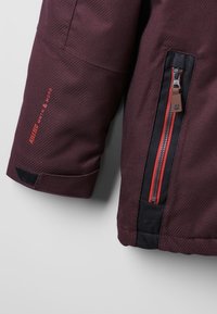 Bordeauxjack met mouw met rode tekst "KALIBER MANTM & MORE", een verticale ritszak in zwart en rood, en een verstelbare manchette met zwarte klittenband.