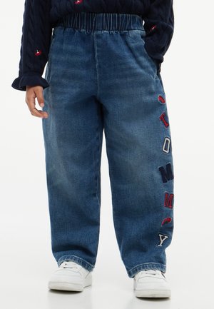 Enfant portant un jean bleu ample avec une taille élastique et des lettres brodées sur une jambe, associé à des baskets blanches et un pull bleu marine.