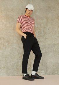 Rot-weiß gestreiftes T-Shirt, schwarze Hose mit Falten, weiße Mütze und schwarze Sneakers mit pinkem Akzent, stehend vor einem Betonhintergrund.