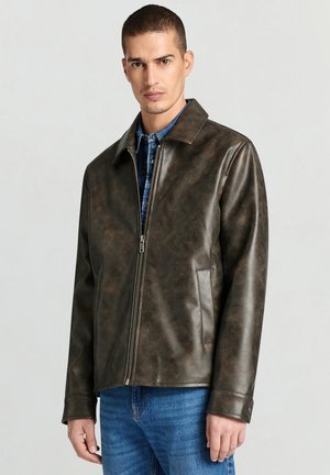 Mann trägt braune Lederjacke mit Reißverschluss über blauem Jeanshemd und Jeans, steht vor hellem, einfarbigem Hintergrund.