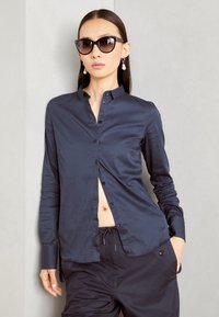 Camicia navy con bottoni in tessuto liscio, dotata di colletto e spacco sul davanti, abbinata a pantaloni scuri larghi.