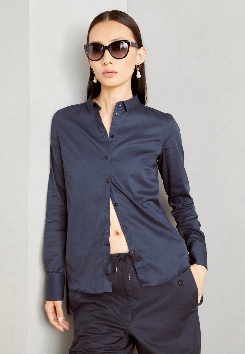 Camicia navy con bottoni in tessuto liscio, dotata di colletto e spacco sul davanti, abbinata a pantaloni scuri larghi.