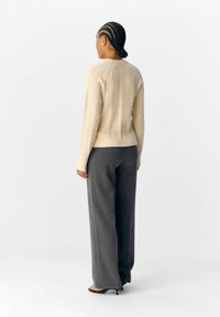 Beige kabelstickad tröja med långa ärmar, rund halsringning och ribbad nederkant, kombinerad med löst sittande grå byxor och svarta klackskor.