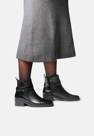 Femme portant des bottines en cuir noir à talons bas et boucles, associées à des collants noirs transparents et une jupe en laine grise.