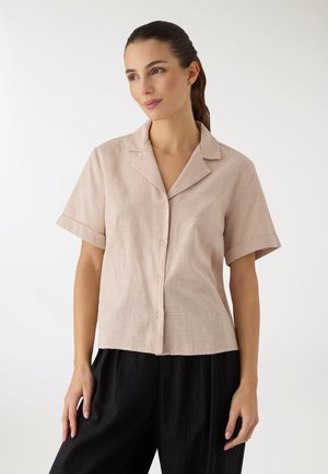 Donna con capelli scuri legati in una coda di cavallo, indossa una camicia beige a maniche corte con bottoni e pantaloni neri larghi, in piedi davanti a uno sfondo bianco uniforme.