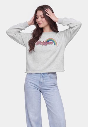 Ung kvinde med langt brunt hår iført en lysegrå sweatshirt med en regnbue og teksten "Rainbow", poserende med hænderne på hovedet.