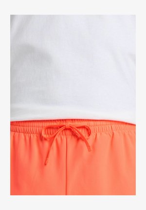 T-shirt en coton blanc au-dessus de shorts à taille élastique orange vif avec cordon de serrage. Les shorts présentent un bandeau élastique froncé et un nœud.