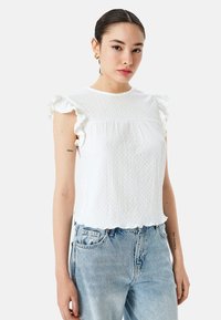 Top blanco texturizado con mangas con volante, escote redondeado y dobladillo con forma de concha. Combinado con jeans de mezclilla azul claro.