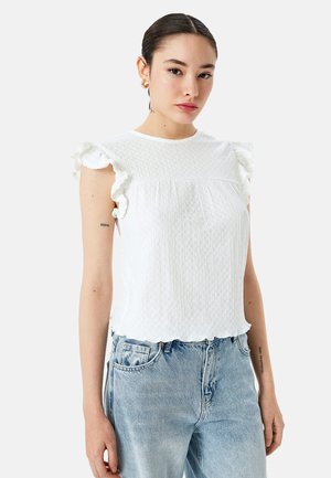 Haut blanc texturé avec des manches à volants, encolure ronde et ourlet festonné. Associé à un jean en denim bleu clair.
