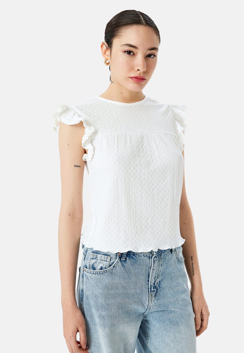Top blanco texturizado con mangas con volante, escote redondeado y dobladillo con forma de concha. Combinado con jeans de mezclilla azul claro.