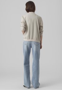Persoon met schouderlang bruin haar, gekleed in een beige jack en lichtblauwe wijde jeans, staand met het gezicht naar een effen grijze achtergrond.