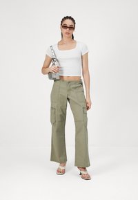 Λευκό εφαρμοστό crop top με τετράγωνη λαιμόκοψη, ελαιόχαρτα cargo παντελόνια σε ελαιώδες πράσινο χρώμα με πλαϊνές τσέπες και γήινες γόβες. Τα αξεσουάρ περιλαμβάνουν μια μικρή γκρίζα τσάντα.