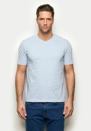 Mann mit kurzen braunen Haaren, der ein hellblaues V-Ausschnitt-T-Shirt und blaue Jeans trägt, steht vor einem schlichten weißen Hintergrund.