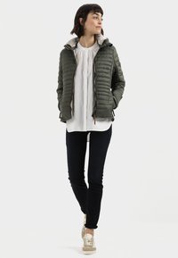 Olijfgroene gewatteerde jacket met een ritssluiting aan de voorkant, ribbelstructuur en een lichte binnenvoering. In combinatie met een wit shirt en donkere jeans.