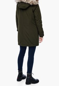 Parka de color verde oliva con cuello de piel sintética, tela texturizada, longitud por debajo de la cadera, combinada con jeans ajustados azul oscuro y botas de tobillo negras.
