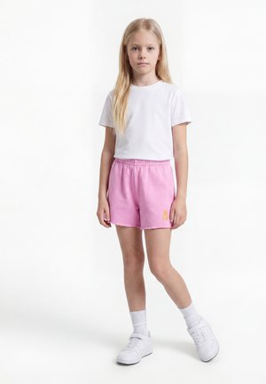 Jeune fille aux longs cheveux blonds se tient debout, portant un t-shirt blanc, un short rose, des chaussettes blanches et des baskets blanches sur fond blanc.