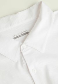Witte collshirt gemaakt van lichtgewicht stof, met een gladde textuur, subtiele glans en een knoopsluiting aan de voorkant met een merklabel.