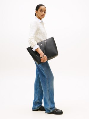 Black leather tote bag with silver hardware, featuring a smooth texture and a structured shape, held by a person wearing a cream cable-knit sweater.