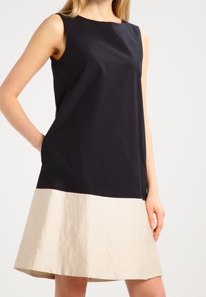 Mouwloze jurk met een zwart topje en een beige rok, gedragen door een persoon met één hand achter de rug.