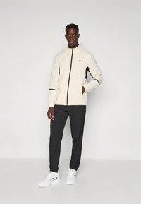 Casaco bege claro com fecho éclair, com detalhes em preto, combinado com calças escuras e ténis brancos. O casaco apresenta um logótipo no peito e bolsos com fecho de correr.