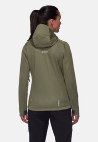 Olivegrüne Softshell-Jacke mit figurbetontem Design, Kapuze, Reißverschlusstaschen und Logos auf dem Rücken und dem unteren Saum. Strukturierter, wetterbeständiger Stoff.