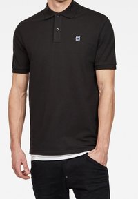 Musta pokeripaita, jossa on kaulus, kaksipainikeinen napitus ja pieni logo vasemmassa rinnassa; valmistettu sileästä, pehmeästä kankaasta.