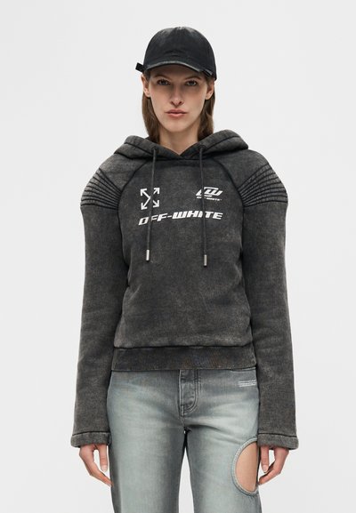 Γυναίκα που φοράει μαύρο καπέλο, γκρι σκούρο hoodie Off-White με επενδυμένους ώμους και γκρι τζιν με μεγάλο άνοιγμα στο γόνατο.